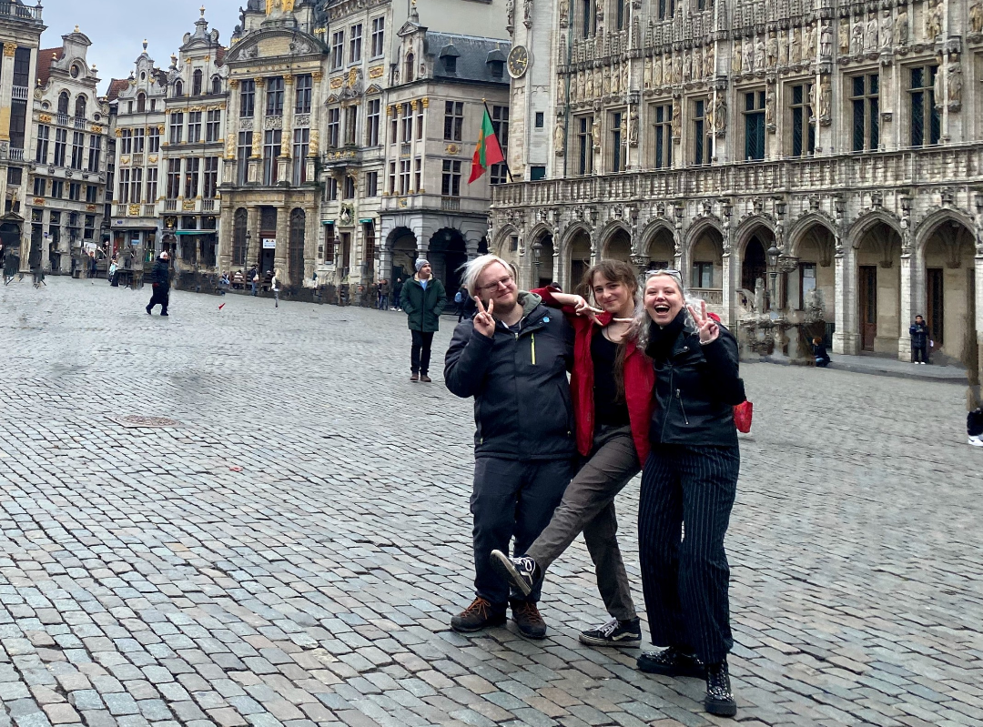 A picture of 3 people posing for a photo together doing peace sign hands. They are in an open square with old buildings around them