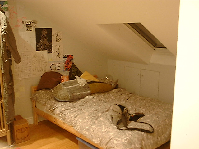 A photo of a bedroom, the quality is grainy and the colour is slightly yellow, in the room is a bed with a large cluster of posters behind it, cushions and a large stuffed shark rest on top of it.